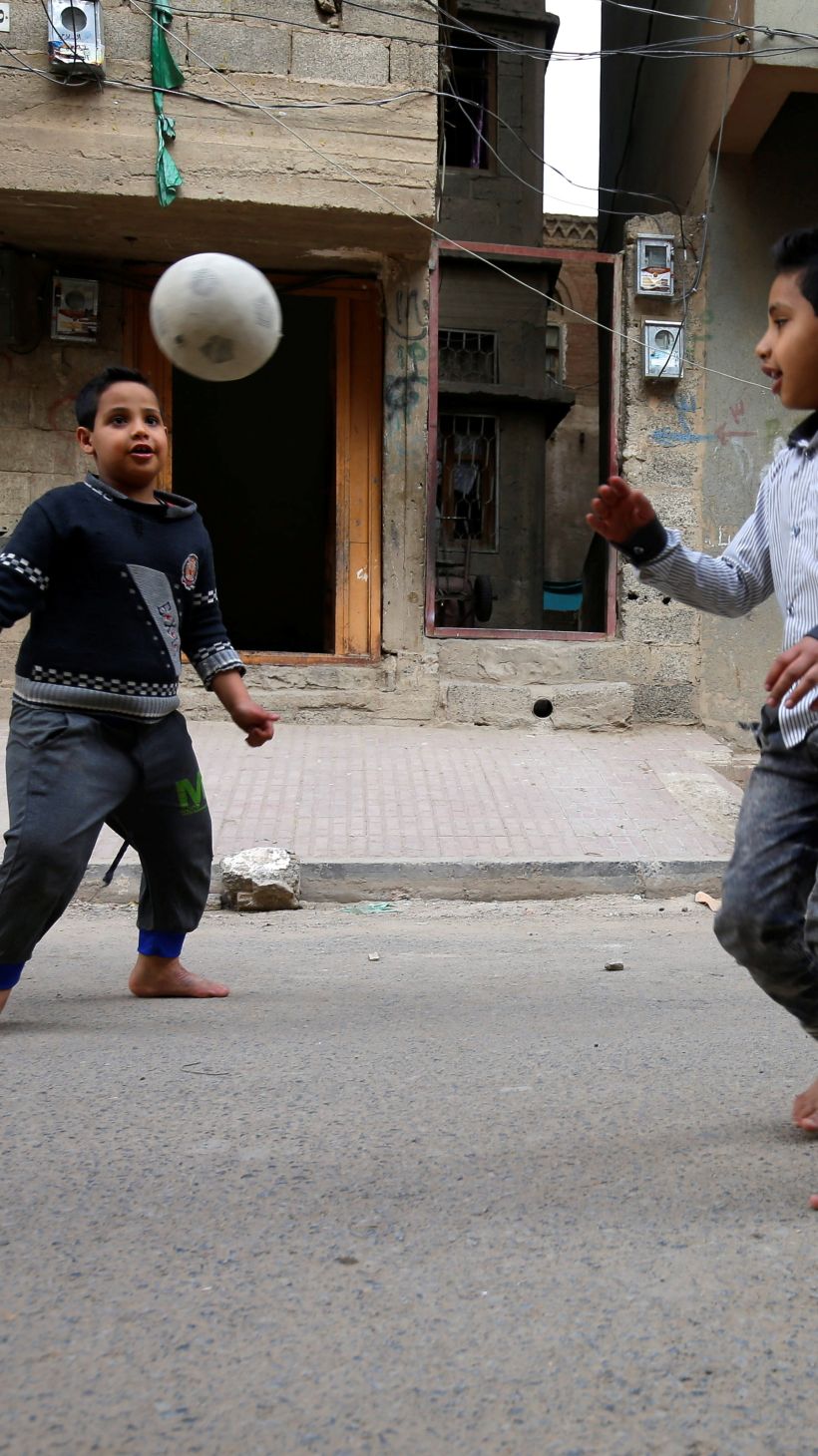 A photo of two kids playing football in the street, outside of a building with a red car in front of it.