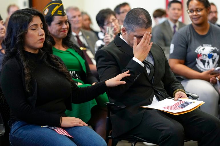 Mata in a black suit, sitting near his wife, has his head in his hand looking emotional as his wife rests her hand on his shoulder in comfort