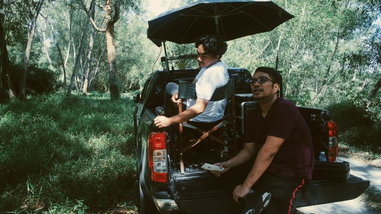 Amir Muhammad and a colleague seated at the back of a truck on the set of his film Roh. They are in a forested area.