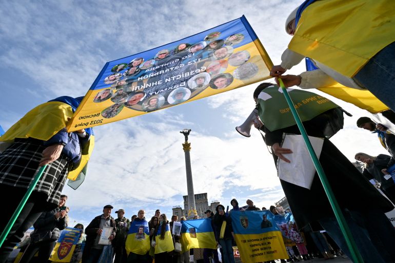 A protest in central Kyiv by families of soldiers missing in the war. They are holding a blue and yellow flag with pictures of their loved ones. A monument can be seen behind.