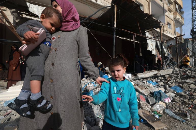 a mother holding small children walks past a destroyed building