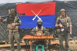Azad, a US national, (left) and an unnamed British fighter (right) with the PDF Zoland. The two men are standing either side of a desk where another fighter is sitting with weapons on display. There is a flag behind them. The men are all masked.
