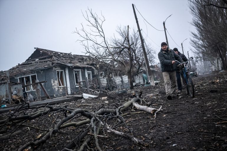 two men wheel a bicycle past a destroyed building