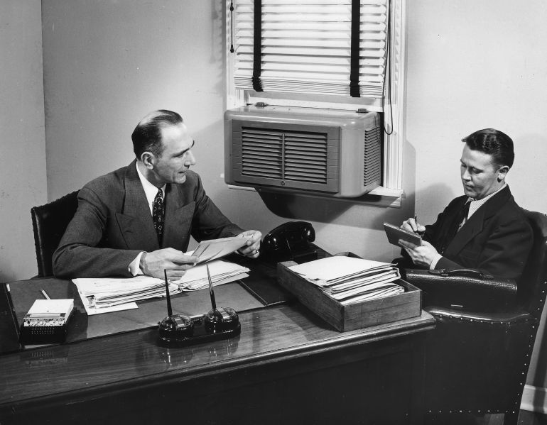An air conditioner is mounted in the window of an office, circa 1955