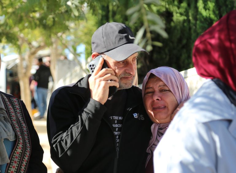 Raed wears a baseball cap. he's middle-aged with frey stubble. he's on his phone and has an arm around Ibtisam who is crying