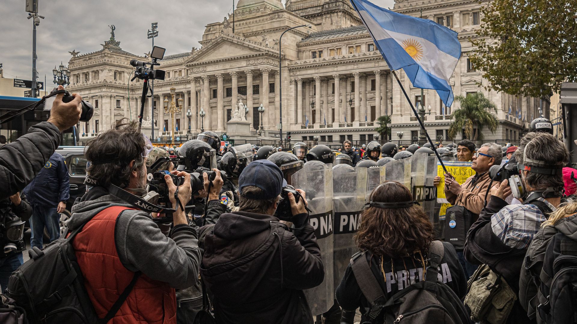 A wall of police officers in riot gear, with large plastic shields, stands near the Congress building while journalists take photographs and protesters demonstrate.